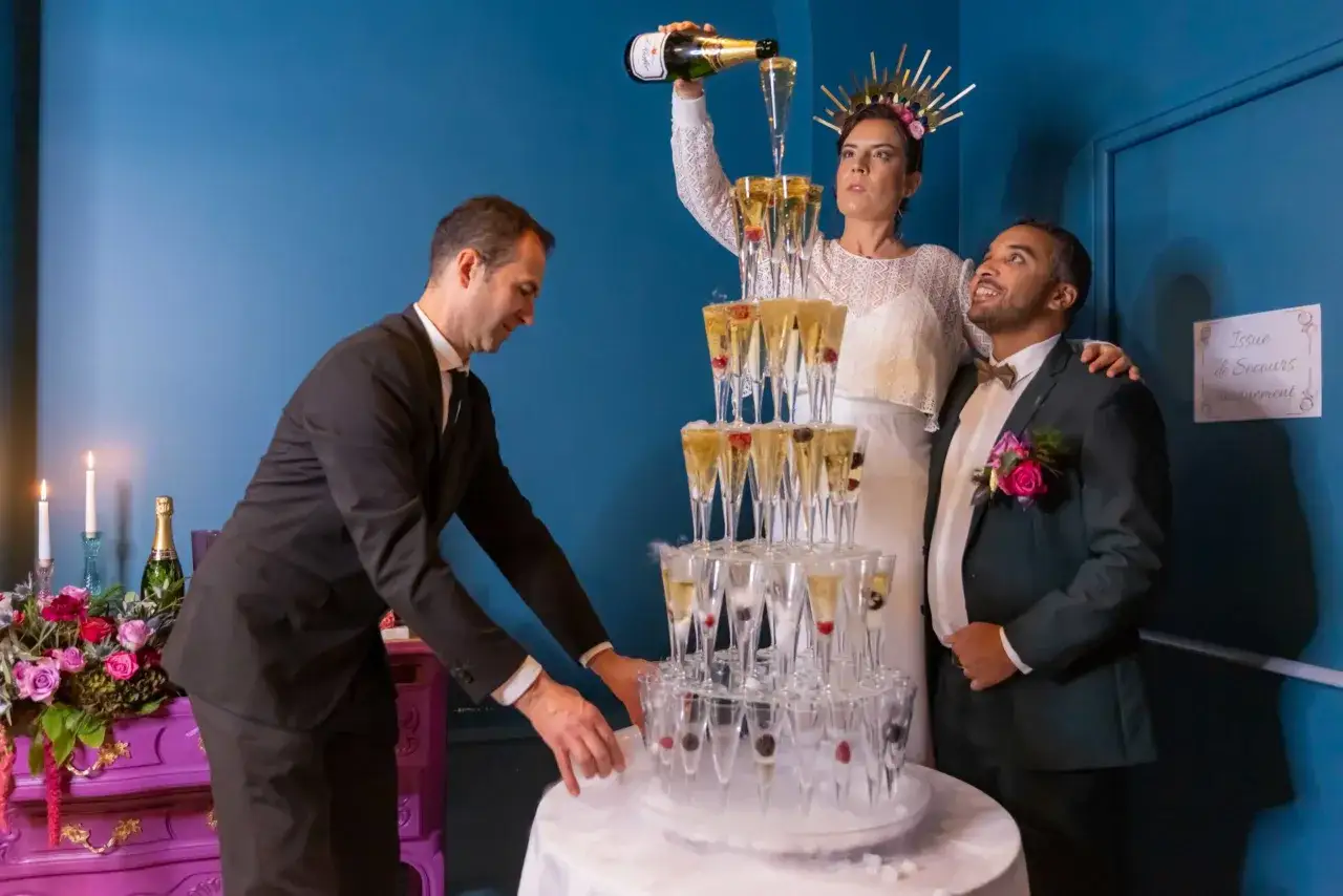 Un homme verse du champagne dans une pyramide de verres pour un vin d'honneur pour 100 personnes. La mariée, couronnée, et le marié sourient.