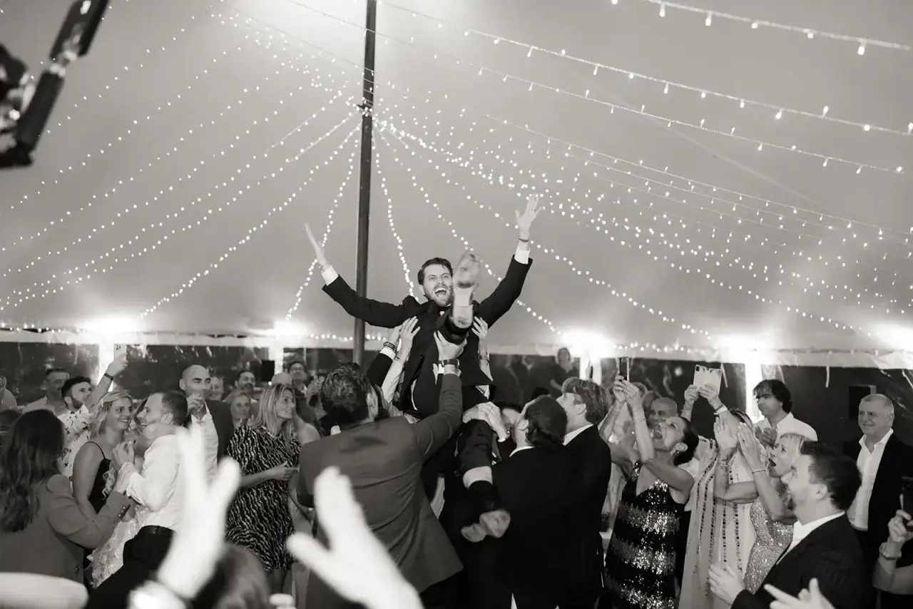 Mariage sous guirlandes lumineuses. Un homme est porté par la foule, joyeux, comme sur une playlist mariage toute faite.