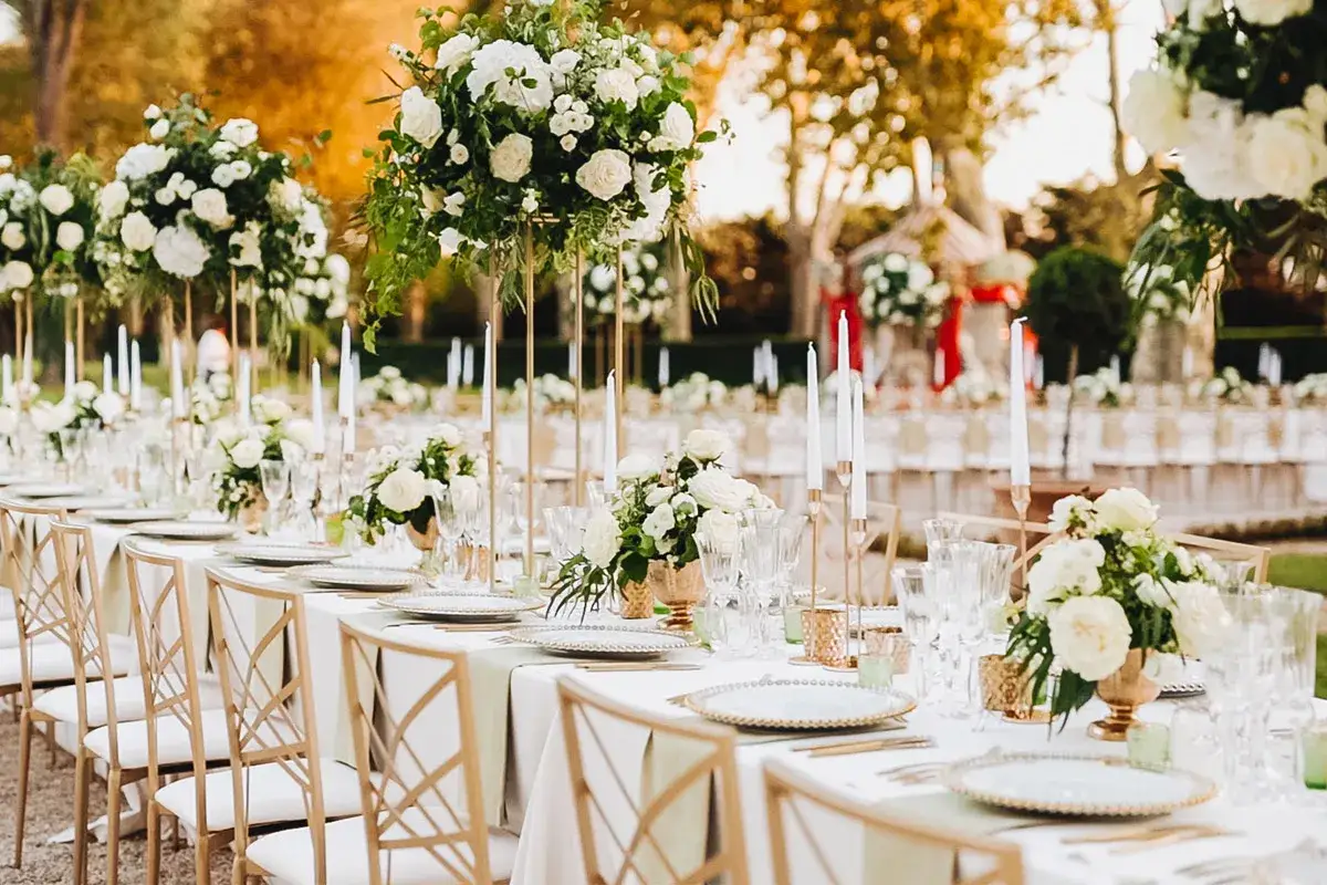 Disposition table mariage élégante en extérieur, avec des centres de table floraux blancs, des bougies et des chaises dorées.