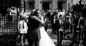Un couple s'embrasse devant une église, entouré d'invités. Un jeune garçon tient un bouquet. Ce moment est le fruit d'un savant **rétroplanning mariage**.