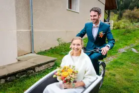Un jeune couple souriant sur un vélo cargo, la mariée tenant un bouquet. Une idée originale pour un mariage avec un petit budget.