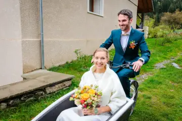 Un jeune couple souriant sur un vélo cargo, la mariée tenant un bouquet. Une idée originale pour un mariage avec un petit budget.