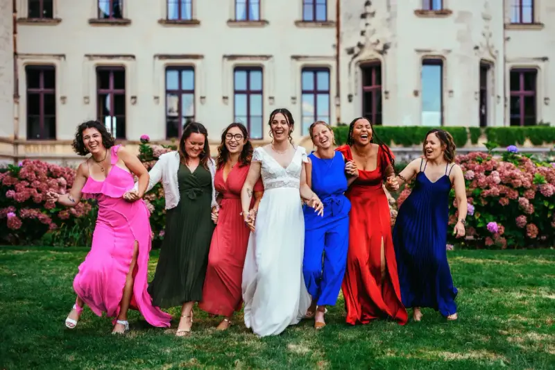 Un groupe de femmes rit joyeusement devant un château. La mariée en blanc est entourée de ses demoiselles d'honneur en robes colorées. La différence entre témoin et demoiselle d'honneur est subtile, mais leur complicité est évidente.